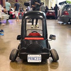 a small black car sitting on top of a hard wood floor in a building with lots of people around it