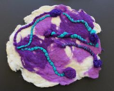 a purple and white piece of art with beads hanging from it's sides on a black surface