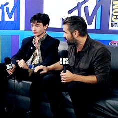 two men sitting on a couch with microphones in their hands and one man standing up