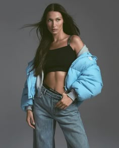 a woman with long hair wearing a black top and blue puffy jacket, standing in front of a gray background