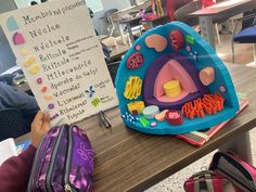 a child is sitting at a table with a sign in front of her and other children's backpacks