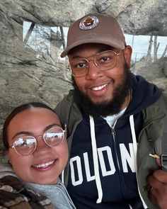 a man and woman taking a selfie in front of a rock wall with trees