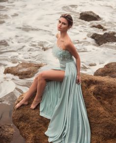 a woman sitting on top of a rock next to the ocean wearing a blue dress