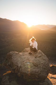 a woman sitting on top of a rock with the sun setting in the distance behind her