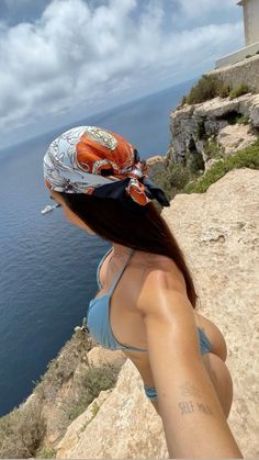 a woman standing on top of a cliff next to the ocean wearing a bandana