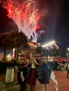 three women pose for a photo while fireworks go off in the sky behind them at night