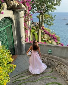 a woman in a pink dress is walking down some steps by the water and flowers