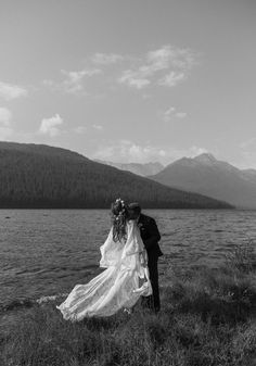 a bride and groom are standing by the water in black and white, with mountains in the background