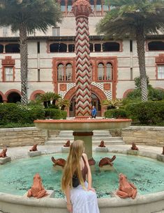 a woman standing in front of a fountain with palm trees around it and a building behind her