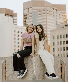 two young women sitting on the edge of a cement wall in front of tall buildings