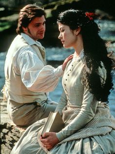 a man and woman sitting next to each other on a rock near the water with trees in the background