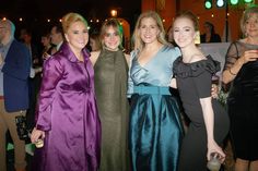 four women in evening dresses posing for the camera at an event with people standing around