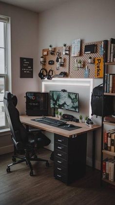 a desk with a computer on it in front of a window and bookshelf