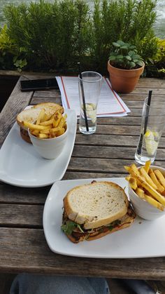 two plates with sandwiches and french fries on them sitting on a wooden table near water