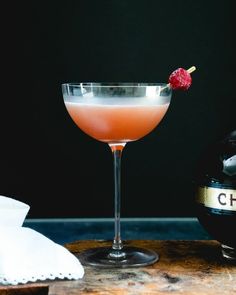 a pink cocktail with a raspberry garnish on the rim in front of a bottle