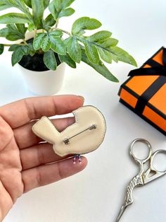 a hand holding a white heart shaped brooch next to a pair of scissors and a potted plant