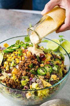 a person pouring dressing into a salad in a glass bowl with lettuce, corn and other vegetables