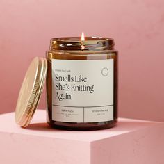 a candle that is sitting on top of a pink block with the words smells like she's knitting again