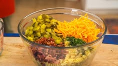 a glass bowl filled with pasta, cheese and pickles on top of a wooden table