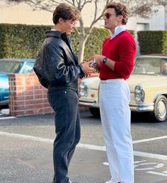 two people standing in the middle of a parking lot talking to each other with cars behind them