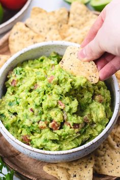 someone dipping guacamole into a bowl with tortilla chips and tomatoes in the background