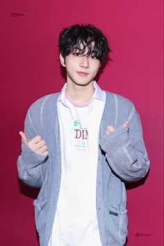 a young man with black hair wearing a white shirt and grey cardigan jacket standing in front of a pink wall