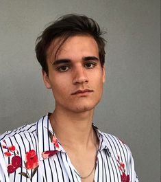 a young man wearing a striped shirt with red flowers on the collar and chest is looking at the camera