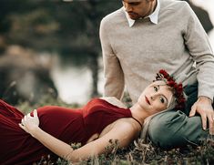 a man standing next to a woman in a red dress laying on the ground near water