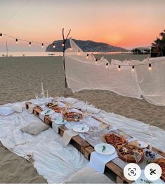 a table set up on the beach at sunset with food and drinks laid out in front of it