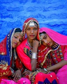 three women dressed in bright colored clothing sitting next to each other on the ground with their heads together