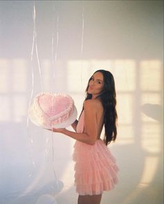 a woman in a pink dress holding a heart shaped cake with balloons attached to it