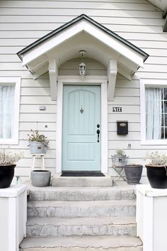 the front door of a white house with potted plants on the steps and two planters