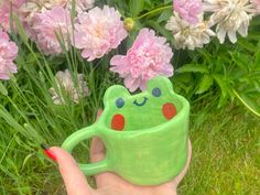 a hand holding a green frog mug in front of pink flowers