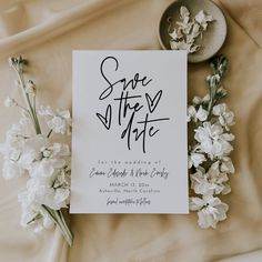 a wedding save the date card and some flowers on a bed with white linens