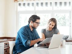 two people sitting at a kitchen counter looking at something on a laptop computer, while the woman is smiling