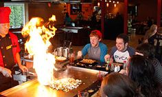 a group of people sitting around a table with food on top of it and flames in the air