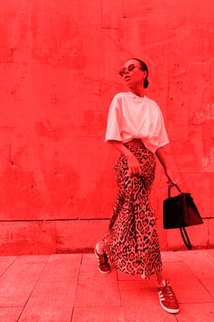 a woman standing in front of a red wall holding a handbag and wearing sunglasses