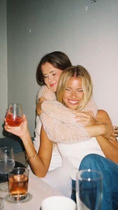 two women hugging each other at a table with wine glasses and drinks in front of them