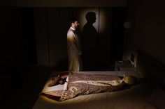 a man standing next to a bed in a dark room with his shadow on the wall