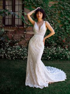 a woman in a white dress standing on grass with her hands behind her head and looking off to the side