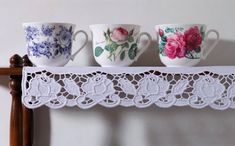 three cups are sitting on a shelf with lacy doily around the cup holders, which have roses and leaves painted on them