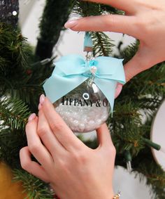 two hands holding an ornament in front of a christmas tree with the words tiffany & co on it