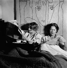 two women sitting on a bed looking at something in front of the woman is reading