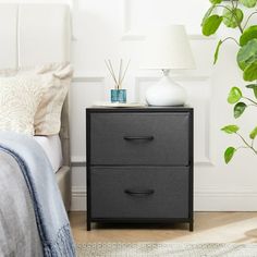 a nightstand with two drawers next to a bed and a plant on the side table