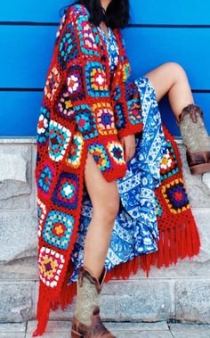 a woman wearing cowboy boots and a colorful crochet blanket sits on steps in front of a blue wall