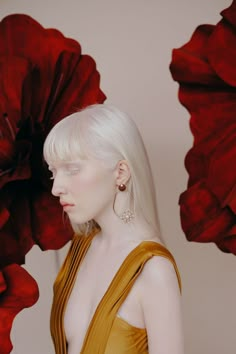 a woman with blonde hair and earrings standing in front of large red flower petals, looking off to the side