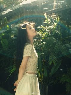 a woman standing in front of some plants and looking up at the sky with her eyes closed