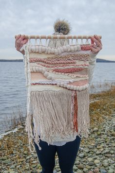 a woman standing on rocks holding up a woven wall hanging over the water's edge