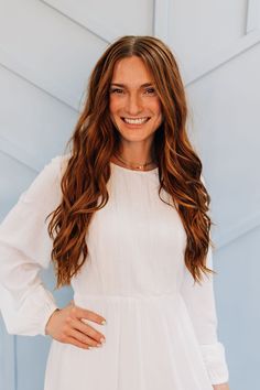 a woman with long brown hair wearing a white dress and smiling at the camera while standing in front of a blue wall