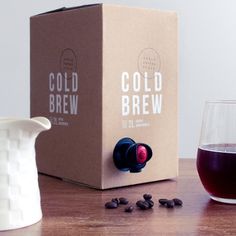 a glass of cold brew next to a box and pitcher on a table with coffee beans